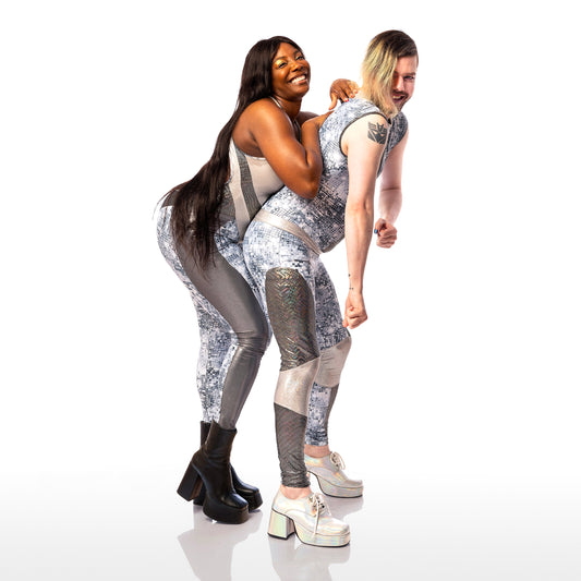 Disco Diva Leggings, a handmade-to-order piece in disco ball and silver foil print spandex. The leggings feature a double layer high waist with light silver knee panels and granite foil curved leg panels. Models Max stands side on to the camera and leans forward. Model Roslyne stands behind Max leaning on their back. Roslyne wears a matching catsuit. 