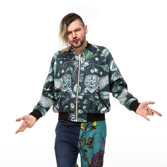 Extinct Bomber Jacket, a handmade-to-order piece made from cotton satin fabric printed with a blue and green sketched design of lions, dodos and monkeys with skeletons showing. This zip up bomber jacket has raglan sleeves, front welt pockets and ribbed collar, cuffs and waistband. Model Max stands on one foot facing the camera with arms out to the side.  the camera