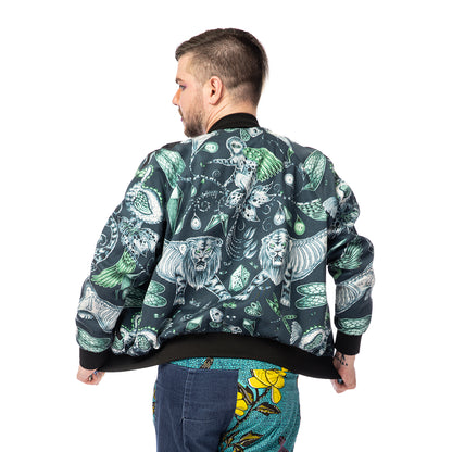 Extinct Bomber Jacket, a handmade-to-order piece made from cotton satin fabric printed with a blue and green sketched design of lions, dodos and monkeys with skeletons showing. This zip up bomber jacket has raglan sleeves, front welt pockets and ribbed collar, cuffs and waistband. Model Max stands with their back to the camera holding out the jacket to the sides. 