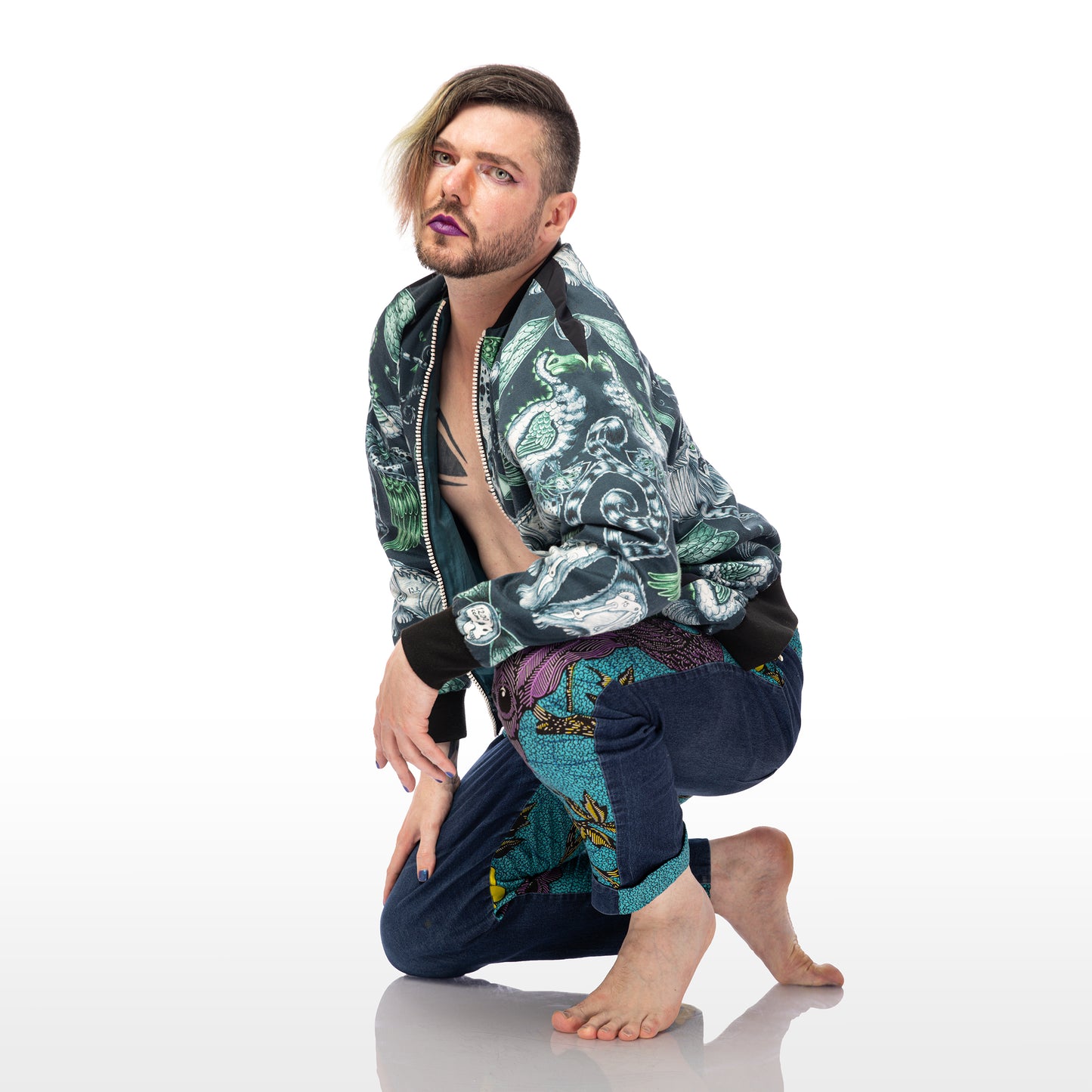 Extinct Bomber Jacket, a handmade-to-order piece made from cotton satin fabric printed with a blue and green sketched design of lions, dodos and monkeys with skeletons showing. This zip up bomber jacket has raglan sleeves, front welt pockets and ribbed collar, cuffs and waistband. Model Max kneels on the floor with one knee and leans on the other. 