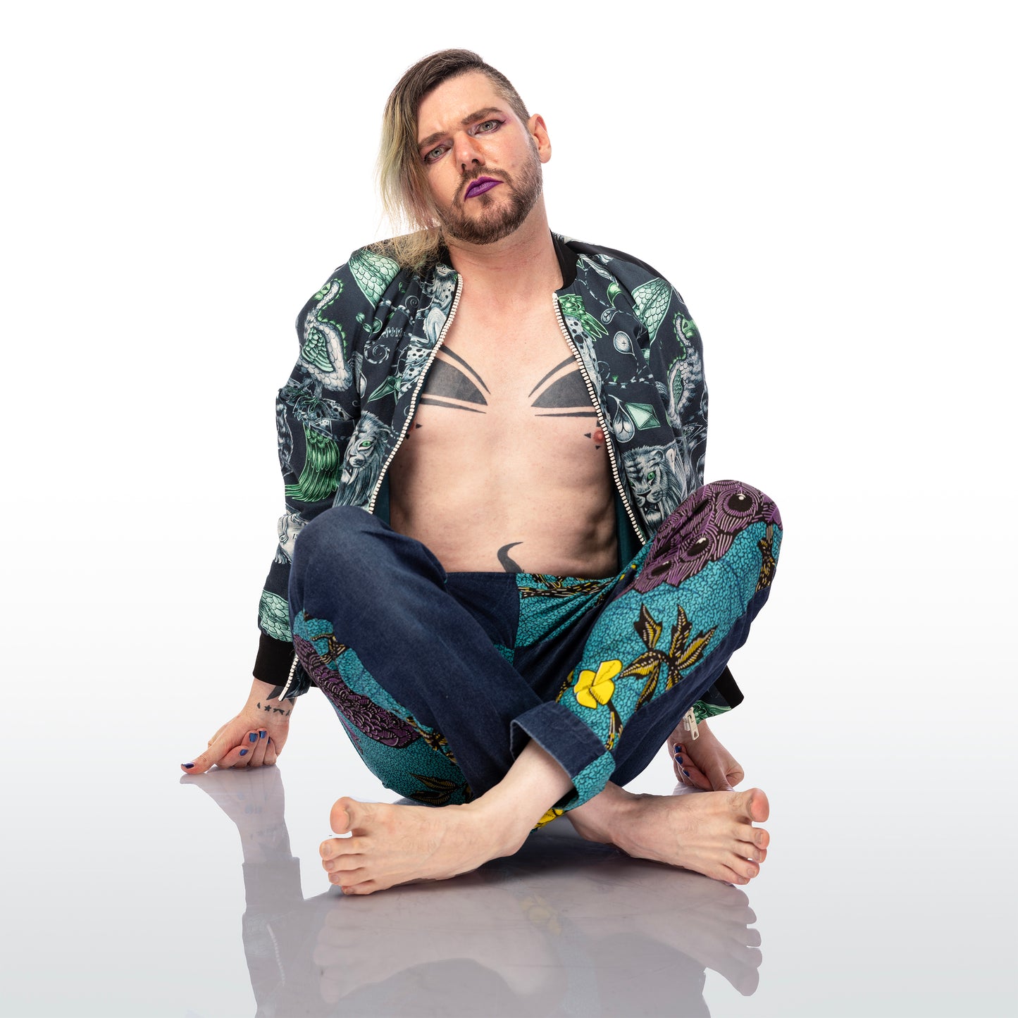 Extinct Bomber Jacket, a handmade-to-order piece made from cotton satin fabric printed with a blue and green sketched design of lions, dodos and monkeys with skeletons showing. This zip up bomber jacket has raglan sleeves, front welt pockets and ribbed collar, cuffs and waistband. Model Max sits on the floor with legs crossed in front of them. 