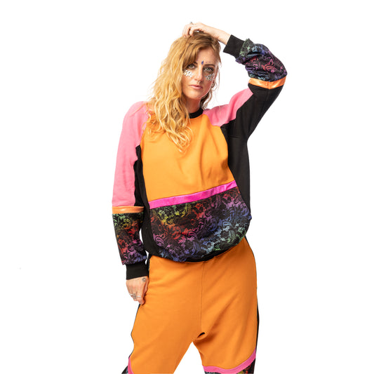 Model Ellie wears the Orange Rainbow Lace Sweater, a handmade-to-order piece in orange, pink and black sweatshirt fleece with rainbow ornamental lace print spandex details. The unisex design features a raglan sleeve, a large printed spandex front pouch pocket, matching lower arm panels and orange and pink foiled stripes. She has long blonde curly hair and gems on her cheeks and stands facing the camera with her hand on her head. 