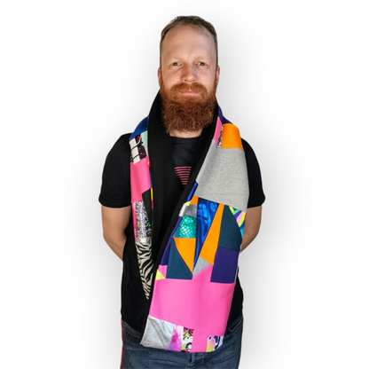 Large circular scarf made from recycled scrap fabrics.  Lined in black fleece and including bright, printed, multi coloured patchwork panels and large colour blocks in grey, pink, purple and blue. Pictured on model hanging loose around neck. . 