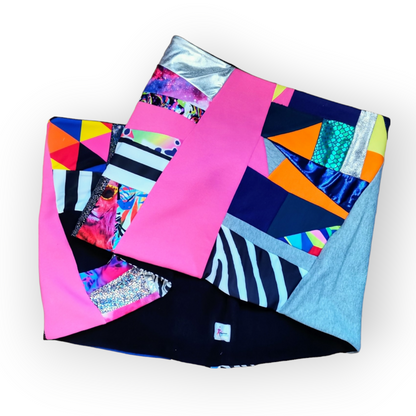 Large circular scarf made from recycled scrap fabrics.  Lined in black fleece and including bright, printed, multi coloured patchwork panels and large colour blocks in grey, pink, purple and blue. Pictured folded to show outer and lining on white background. 