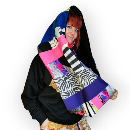 Large circular scarf made from recycled scrap fabrics.  Lined in black fleece and including bright, printed, multi coloured patchwork panels and large colour blocks in grey, pink, purple and blue. Pictured on model hanging loose over head.. . 
