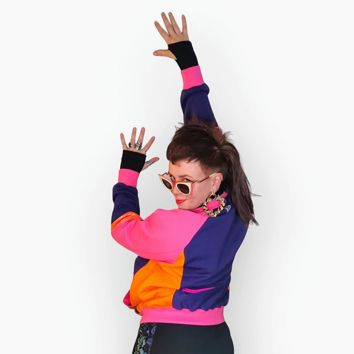 Model Hannah wears Pink Neon Beam Sweater, a limited edition, off-the-peg piece in pink, purple and orange sweatshirt fleece with blue/pink neon beam print spandex details. The unisex design features a raglan sleeve, a large printed spandex front pouch pocket, matching upper arm panels, orange and pink foiled stripes and pink rib cuffs, waistband and neck. They also wear MayHem fabric wrap, heart shaped, large hoop earrings in black and gold and vintage pink and cream sunglasses. Back view. 