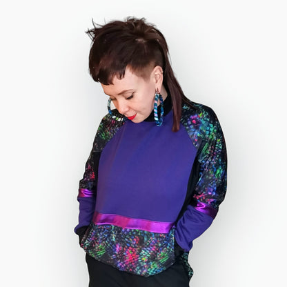 Model Hannah wears the Purple Snakebite Sweater, a limited edition, off-the-peg piece in purple and black sweatshirt fleece with blue/green/pink snakebite print spandex details. The unisex design features a raglan sleeve, a large printed spandex front pouch pocket, matching upper arm panels and pink foiled stripes. They also wear MayHem fabric wrap, large hoop earrings. They are standing facing the camera with their head tilted downwards. 