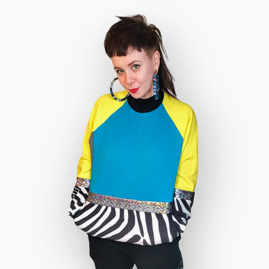 Model Hannah wears Blue Zebra Sweatshirt, a limited edition, off-the-peg piece in blue, yellow and grey sweatshirt fleece with black and white zebra print spandex details. The unisex design features a raglan sleeve, a large printed spandex front pouch pocket, matching upper arm panels, silver hologram foiled stripes and black rib cuffs, waistband and neck. They also wear MayHem fabric wrap large hoop earrings. Front view.