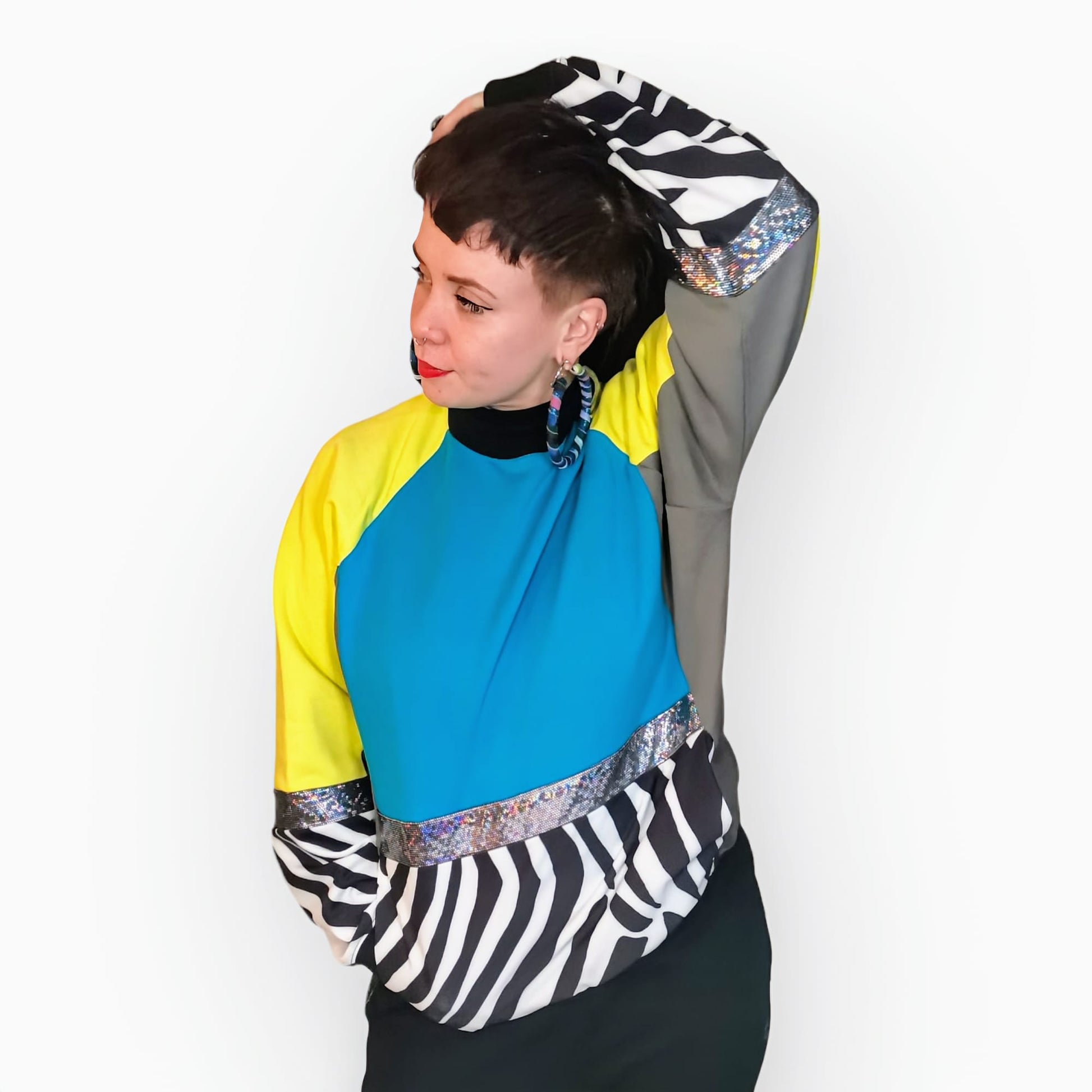 Model Hannah wears Blue Zebra Sweatshirt, a limited edition, off-the-peg piece in blue, yellow and grey sweatshirt fleece with black and white zebra print spandex details. The unisex design features a raglan sleeve, a large printed spandex front pouch pocket, matching upper arm panels, silver hologram foiled stripes and black rib cuffs, waistband and neck. They also wear MayHem fabric wrap large hoop earrings. Font view.