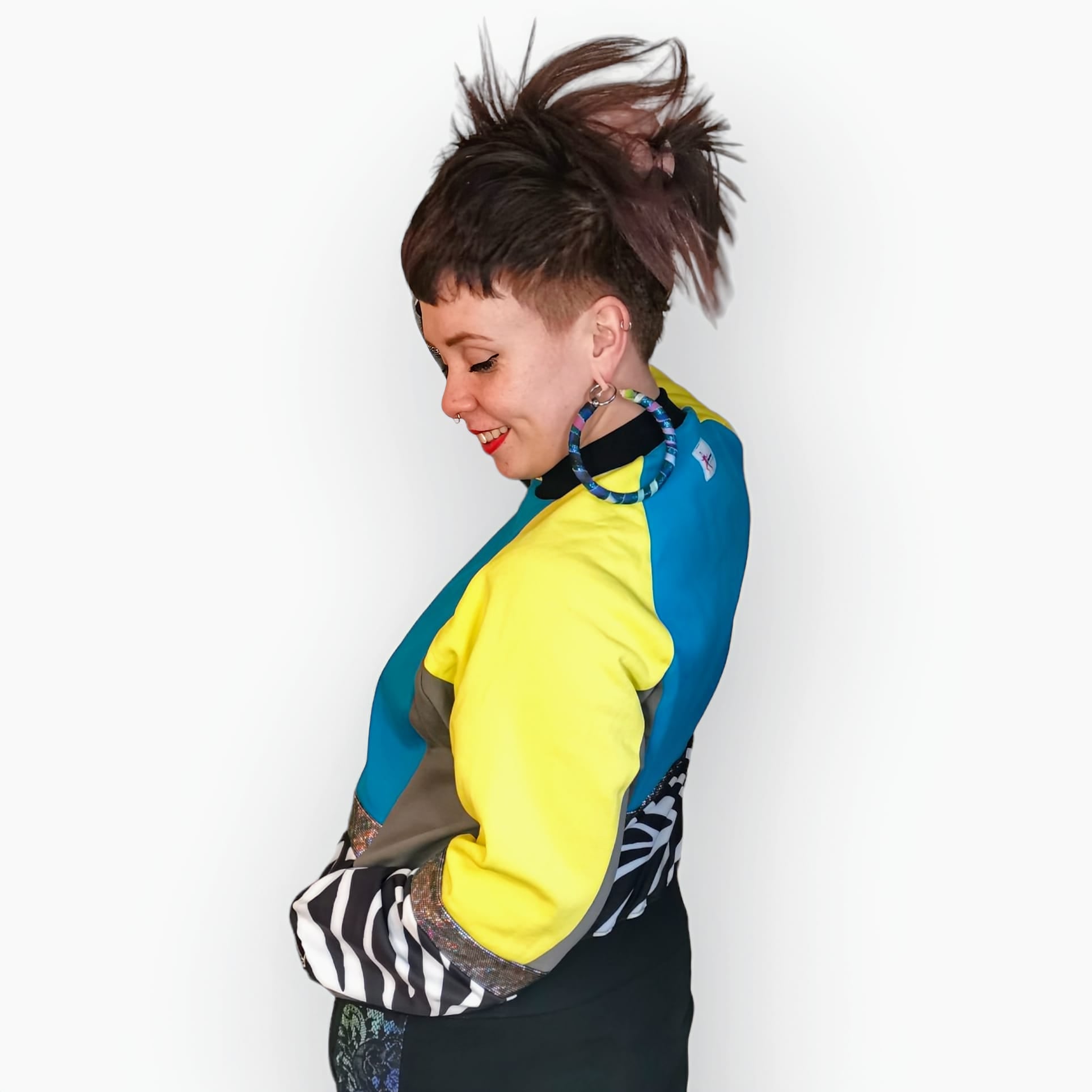 Model Hannah wears Blue Zebra Sweatshirt, a limited edition, off-the-peg piece in blue, yellow and grey sweatshirt fleece with black and white zebra print spandex details. The unisex design features a raglan sleeve, a large printed spandex front pouch pocket, matching upper arm panels, silver hologram foiled stripes and black rib cuffs, waistband and neck. They also wear MayHem fabric wrap large hoop earrings. Side view